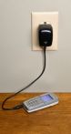 A Cell phone being recharged via a charger that is plugged into an ivory colored outlet that is mounted in a neutral colored wall.